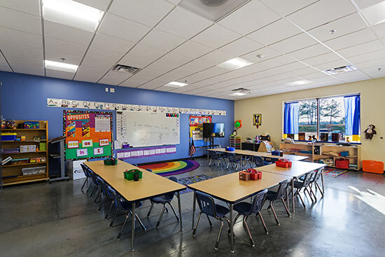 Inside of a classroom with small desks and chairs. poster on the wall, a whiteboard, and shelving of books and toys.