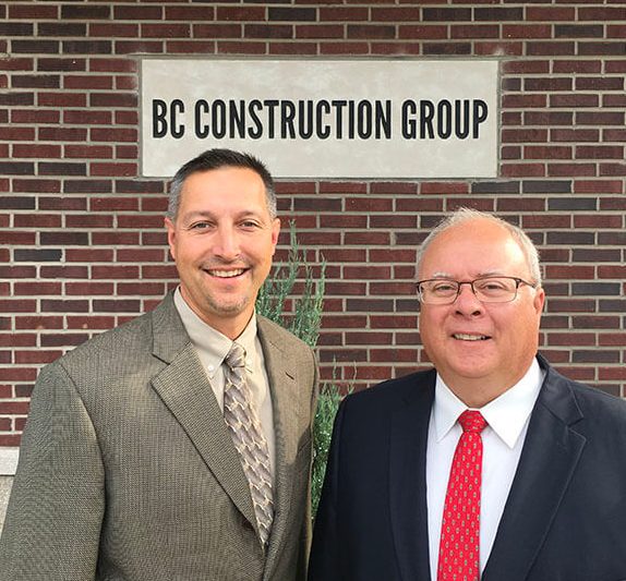 Two male BC Construction Group employees in suits, standing in front of the BC Construction Group sign on building.