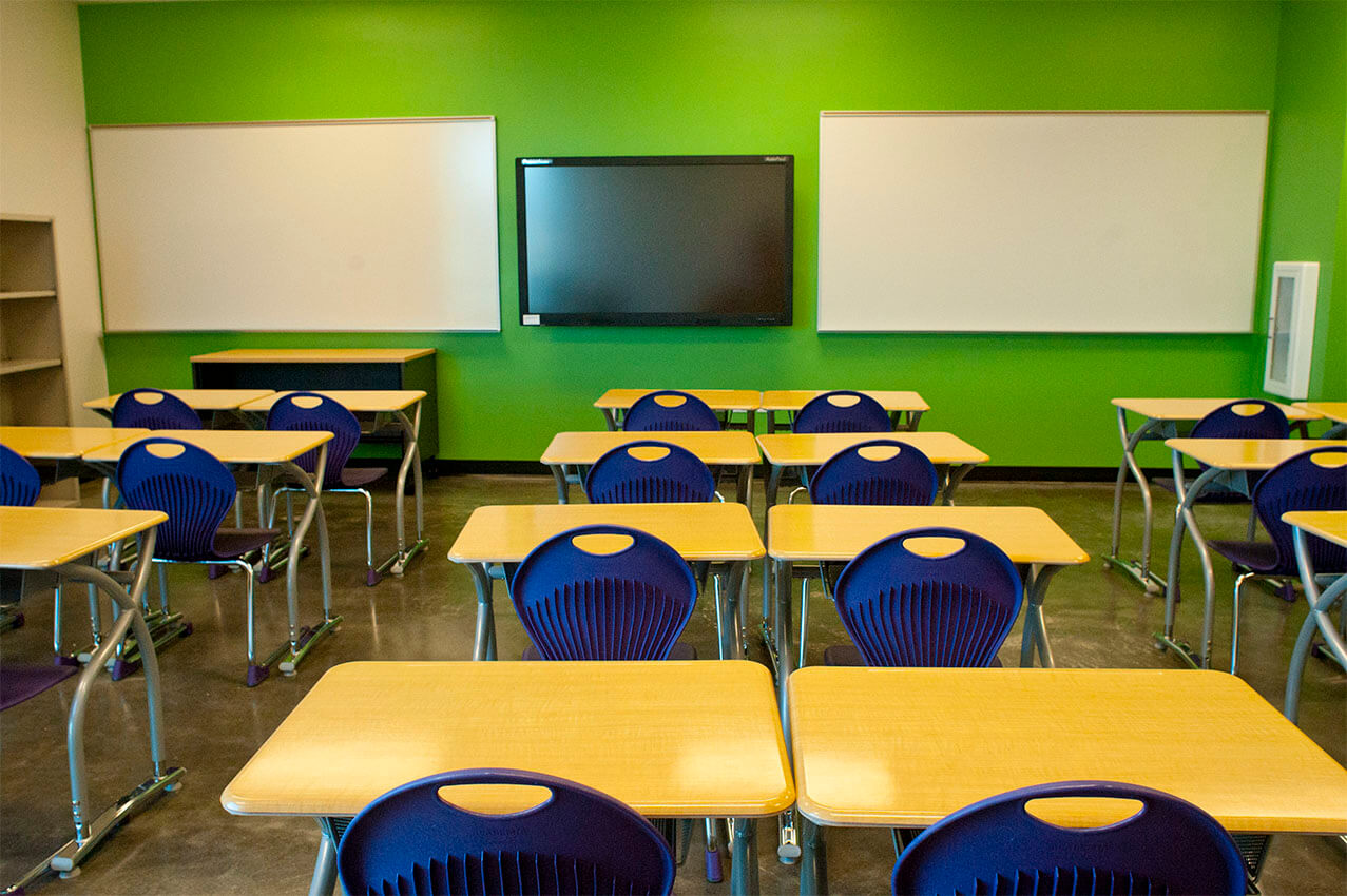 Classroom with green wall, two white boards, and tables with blue chairs.
